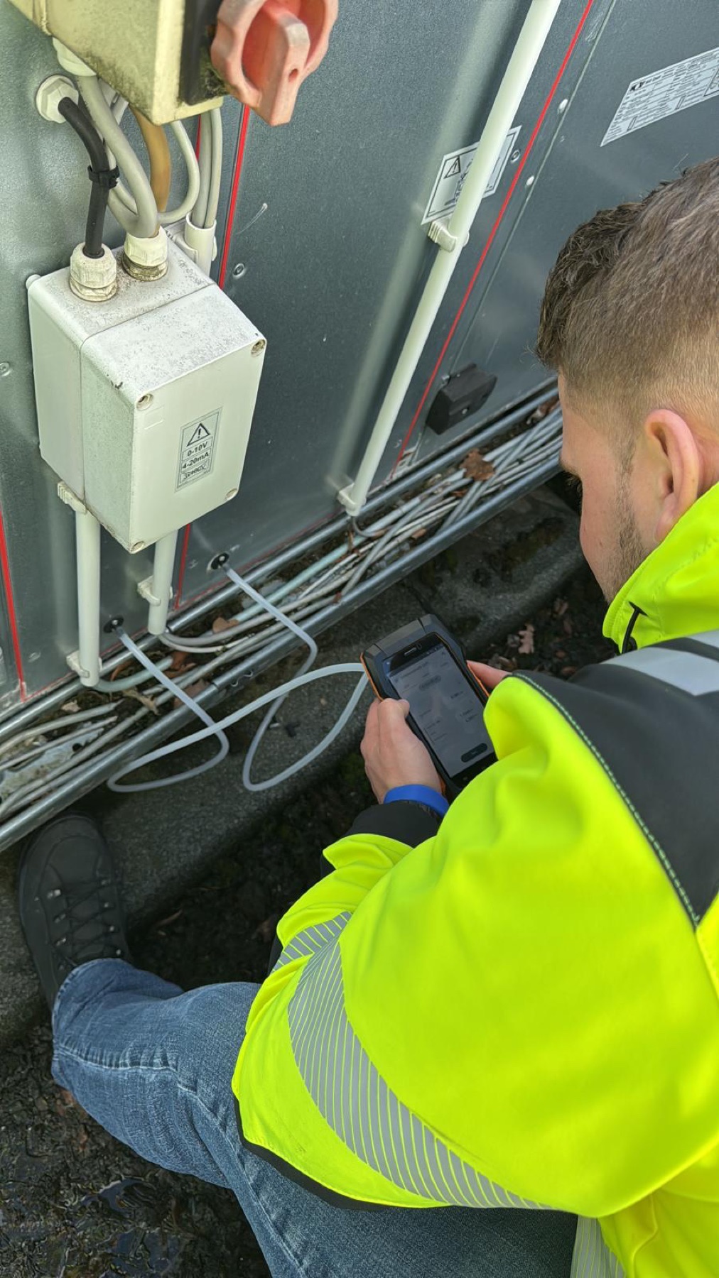 Servicetechniker in gelber Warnschutzjacke schaut auf ein robustes Tablet-Messgerät, im Hintergrund eine geöffnete Lüftungsanlage mit Verkabelung