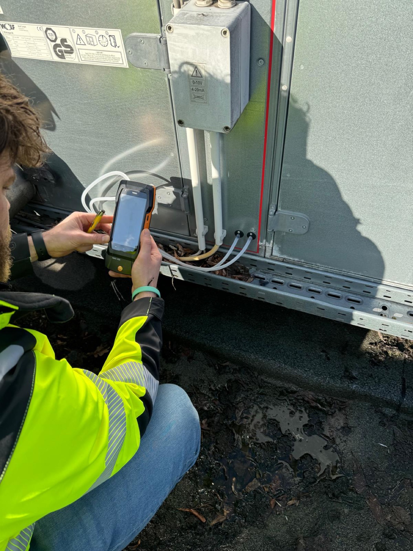 Servicetechniker mit gelber Warnschutzjacke kniet vor einem Lüftungsaggregat und liest mit einem Handheld-Messgerät die Anlagendaten aus